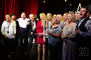 Television makeup artist sydney: group of performers singing together on stage in front of a red curtain