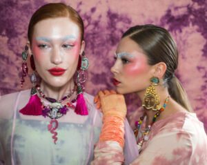 Two models with graphic white and blue eye makeup and red lips, statement jewellery, against a pink backdrop