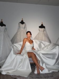 Woman seated on a satin wedding gown train in a bridal boutique, gowns on mannequins behind her