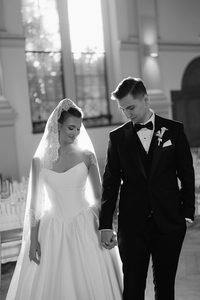 Mobile makeup artist Sydney: bride and groom walking hand in hand down the aisle in black and white, her lace-edged cathedral veil trailing