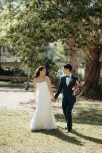 Wedding makeup enquiry Sydney: bride and groom walking hand in hand across the grass beneath an old tree