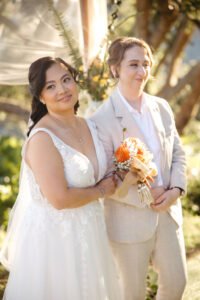 Bridal hair & makeup artist Sydney: two brides after the ceremony, one in a lace V-neck gown, the other in a cream suit with an orange bouquet