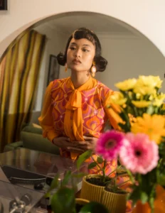 Woman with a vintage finger-wave hairstyle in an orange and pink blouse, styled among flowers